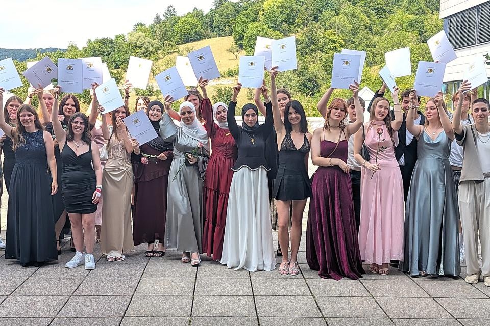 In der Mitte des Bildes sieht man mehrere Personen. Die Personen auf dem Bild halten Zeugnisse in die Luft. Die Personen sind sehr stielvoll Gekleidet. Im Hintergrund ist eine grüne Wiese zu erkennen. Im Hintergrund sind Bäume zu erkennen. Der Boden ist aus grauen Steinfließen.