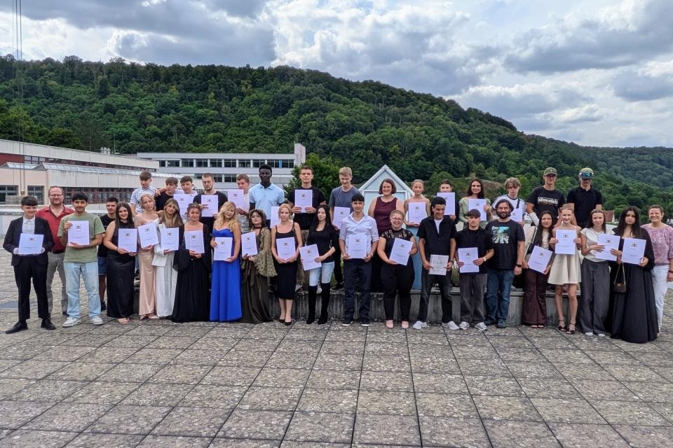 Im Bild sieht man mehrere Personen die schick Angezogen sind. Die Personen auf dem Bild halten alle ein Zeugnis in der Hand. Im Hintergrund sind die Schule und viele Bäume zu sehen. Der Boden besteht aus Steinfließen.