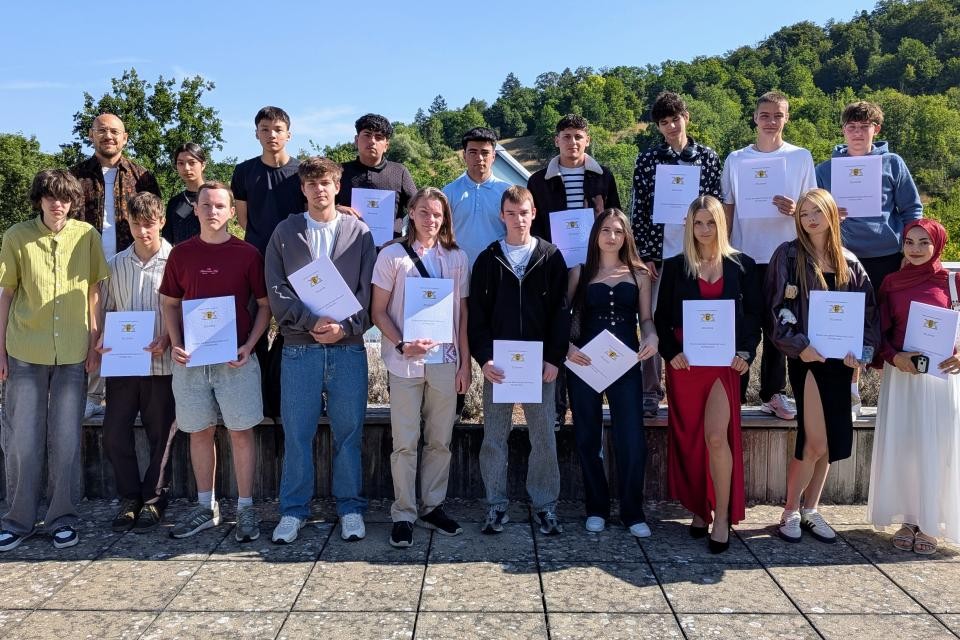 In der Mitte auf dem Bild sieht man junge Menschen die ihr Zeugnis in der Hand halten. Im Hintergrund ist blauer Himmel zu sehen. Im Hintergrund stehen viele Bäume. Der Boden ist aus grauen Steinfließen.