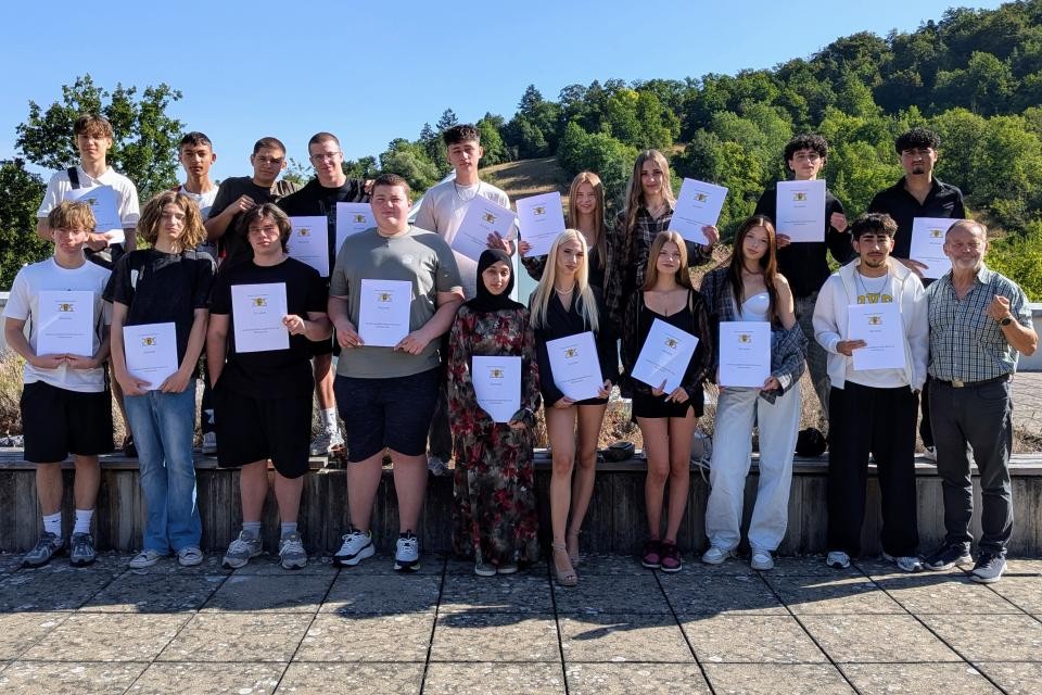 In der Mitte auf dem Bild sieht man junge Menschen die ihr Zeugnis in der Hand halten. Im Hintergrund ist blauer Himmel zu sehen. Im Hintergrund stehen viele Bäume. Der Boden ist aus grauen Steinfließen.