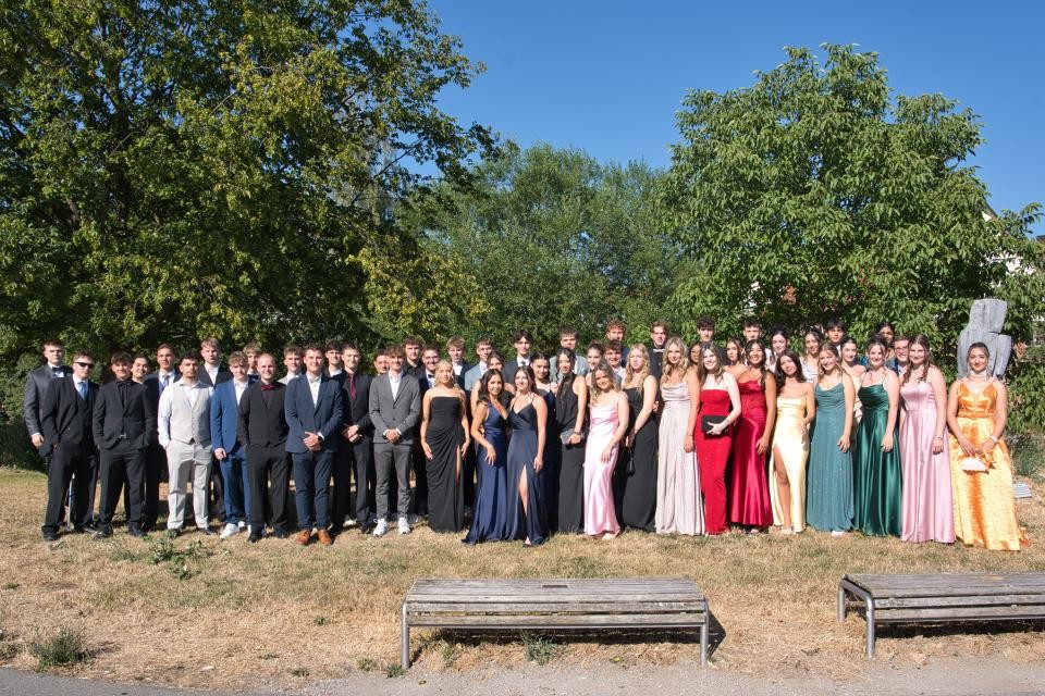 In der Mitte des Bildes sind stielvoll gekleideIn der Mitte des Bildes sind stielvoll gekleidete Personen zu erkennen. Der blaue Himmel ist im Hintergrund zu erkennen. Das Foto wurde auf einer grünen Wiese gemacht. Vor den Personen sind zwei Bänke aus Holz zu sehen. Im Hintergrund sind Bäume zu sehen.