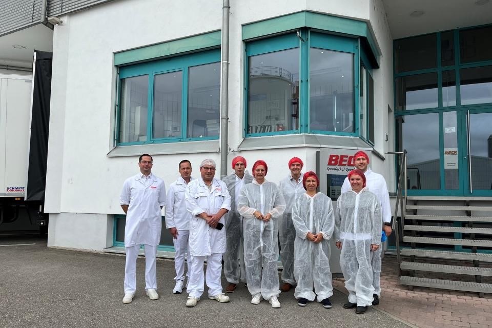 Eine Gruppe von Personen in weiser Schutzkleidung steht vor einem Gebäude mit grünen Fenstern. Rechts hinten ist eine graue Treppe mit einer grünen Eingangstür.