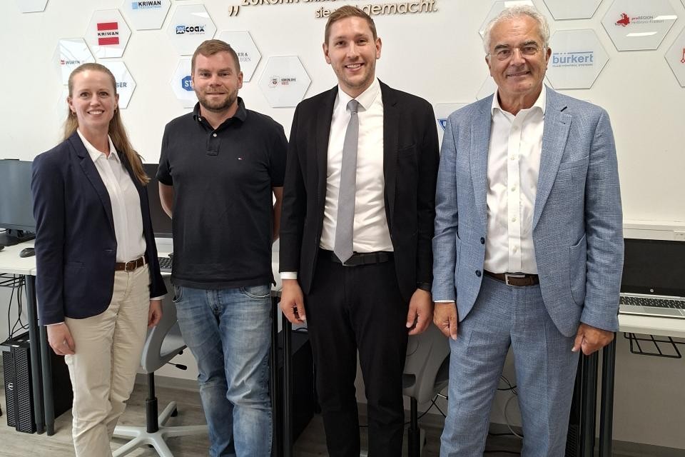 Ein Gruppenbild mit vier Menschen, diese stehen in einem Raum vor einer weißen Wand mit unterschiedlichen Firmenlogos