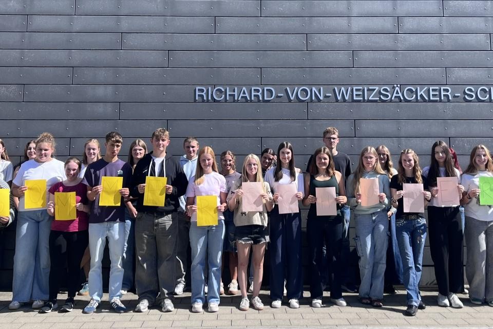 Ein Gruppenbild mehrerer Schüler, sie halten Plakate in der Hand mit der Aufschrift "Sommerschule RWS". Im Hintergrund eine dunkelgraue Wand mit der Aufschrift "Richard-von-Weizsäcker-Schule"