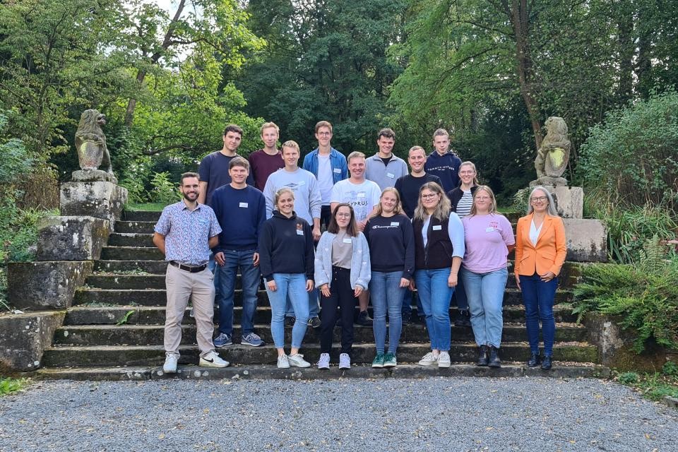 Ein Gruppenbild mehrerer Menschen auf einer großen Steintreppe, an den Seiten der Treppe sitzt links und rechts je ein Löwe aus Stein. Im Hintergrund sind dichte grüne Bäume