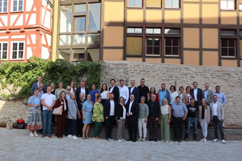 Eine Gruppe von Personen vor einer grauen Steinmauer. Links an der Steinmauer wächst eine grüne Pflanze. Im Hintergrund sind bunte Fachwerkhäuser.