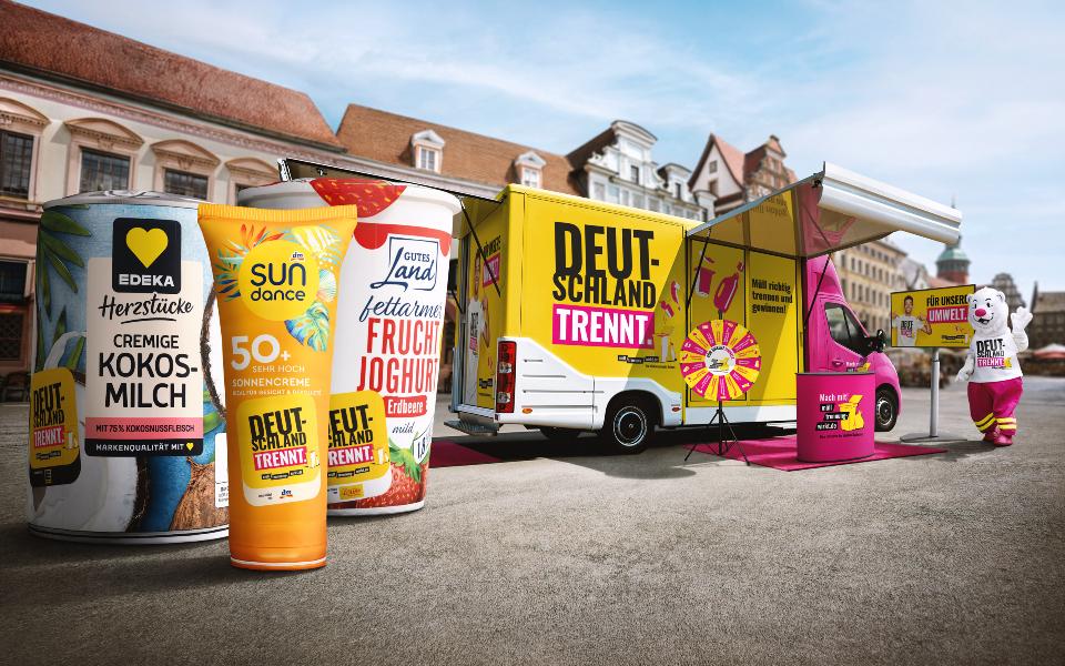 Große bunte XXl-Verpackungen auf grauem Asphalt aufgestellt. Rechts befindet sich der grell gelb-pinke Aktionswagen und Maskottchen "Trenn-Bär".