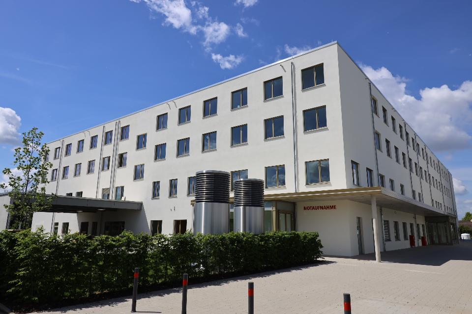 Ein großes weißes Gebäude mit vielen Fenster. Im Vordergrund befindet sich eine grüne Hecke und eine gepflasterte Fläche. Im Hintergrund ist blauer Himmel.