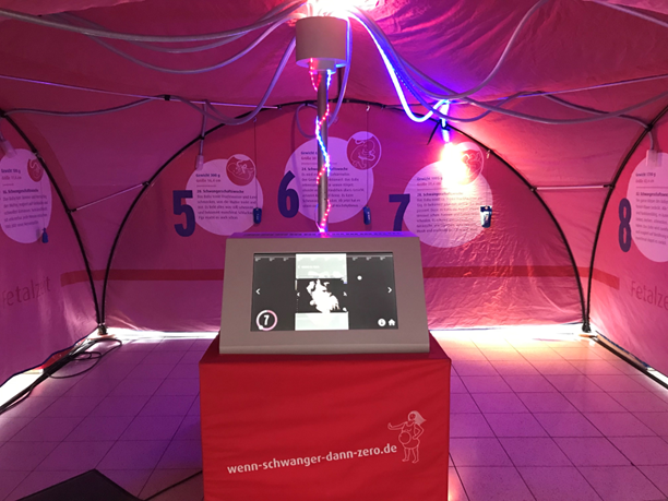 Ein rot beleuchtetes Zelt mit verschiedenen Stationen, in der Mitte befindet sich ein Display