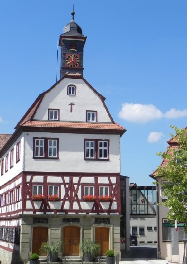 Ein historisches schmales Gebäude. Der erste Stock ist aus hellem Sandstein, darüber folgt ein dunkelrotes Fachwerkkonstrukt. Der dritte Stock ist weiß verputzt. Das Dach verläuft spitz. Auf der Spitze steht ein Turm mit Uhr. Im Erdgeschoss führen drei Holztüren ins innere. Der Himmel ist hellblau mit einzelnen Wolken.
