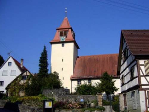 Eine Kirche mit Kirchturm in der Mitte. Rechts und Links befinden sich Wohnhäuser.