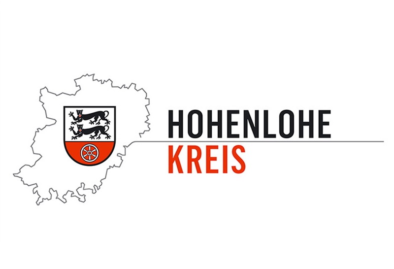 Logo des Landratsamtes Hohenlohekreis. Schwarz, rote Schrift auf weißem Hintergrund: Hohenlohe Kreis. Links Umriss des Landkreises in grau und Wappen des Landkreises: in Silber (Weiß) über erhöhtem rotem Schildfuß, darin ein sechsspeichiges silbernes (weißes) Rad, zwei schreitende, rot bezungte schwarze Leoparden mit untergeschlagenen Schwänzen