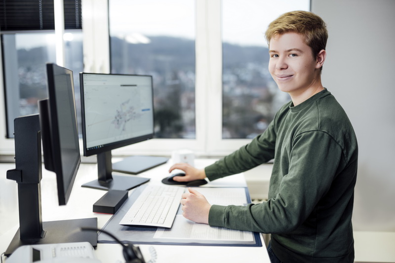 Ein junger Mann am steht am Arbeitsschreibtisch und schaut in die Kamera.