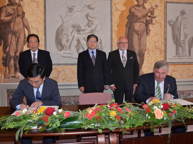 Zwei Personen darunter Landrat Jahn sitzen an einem Tisch mit Blumenschmuck und unterzeichnen die Partnerschaftsurkunde. Im Hintergrund stehen drei weitere Männer.