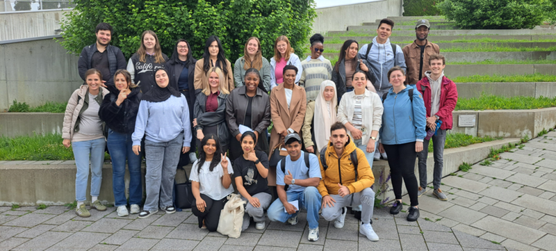Eine Gruppe von Personen steht auf einer gepflasterten Fläche vor einem grünen Busch. Im Hintergrund sind begrünte Treppenstufen.
