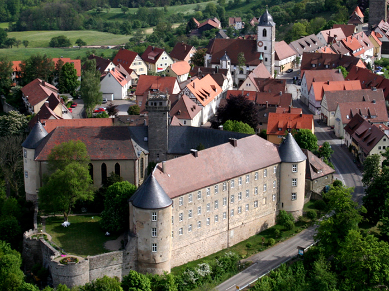 Luftaufnahme: Im Vordergrund steht die Burg mit zwei runden Türmen und einem langen Bau dazwischen. Im Hintergrund stehen Häuser und eine Kirche. Außenherum sind grüne Bäume und Wiesen.