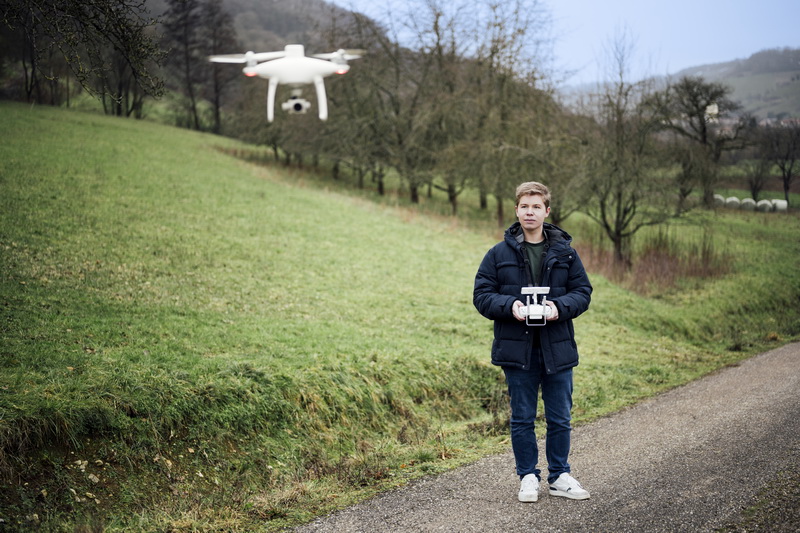 Ein junger Mann steht auf einem Weg neben einer Wiese und steuert eine vor ihm fliegende Drohne.