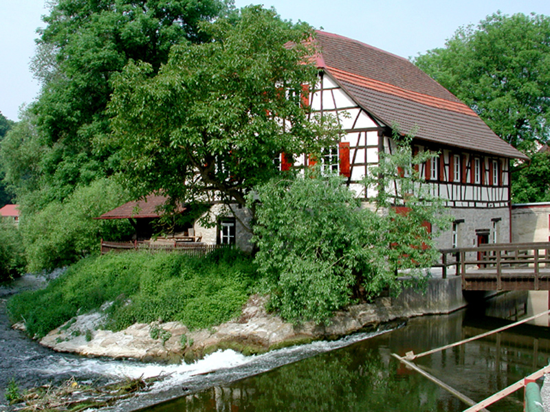 Im Vordergrund ist ein Wehr in der Jagst, einem Fluss, zu sehen. Im Hintergrund ist hinter einem großen Baum mit grünen Blättern eine Fachwerkhaus mit roten Fensterläden zu sehen. Der erste Stock des Hauses ist gemauert. Es handelt sich um die Ölmühle in Dörzbach.